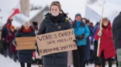 Manifestantes, vestidos com casacos de inverno e com neve ao fundo, carregam cartazes durante uma manifestação em Nuuk, na Groenlândia. À frente, uma manifestante aparece com a boca coberta por fita adesiva
