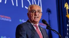Reza Pahlavi usa terno e gravata e fala em um microfone
