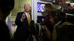 ダークスーツを着て金色のネクタイをしたトランプ氏が、右手を前に出し、人差し指を立てて口を開いている。その前には記者たちがいて、マイクを向けるなどしている