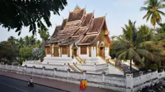 Templo budista em Luang Prabang, pintado de branco e dourado, com três monges andando pela calçada