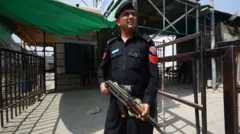 Członek pakistańskich sił bezpieczeństwa, ubrany w ciemny mundur i trzymający karabin automatyczny typu AK‑74, stoi na punkcie kontrolnym.