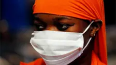 A woman wears a white face mask and covers her hair with an orange fabric.