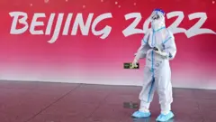 Una persona con un traje de protección junto a una pared con el logo de los juegos olímpicos de invierno.