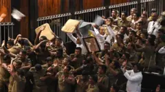 Sri Lankan police escort parliament speaker Karu Jayasuriya (C holding microphone) as aides hold up cushions to protect him and themselves from projectiles thrown by rival legislators in the assembly hall in Colombo on November 16, 2018. - Sri Lanka's parliamentary speaker required police protection as violence returned to the legislature for a second day November 16, with rival factions in the island's constitutional crisis showing no signs of backing down. A group of legislators occupied speaker Karu Jayasuriya's chair for 50 minutes and he was only able to take his seat after he entered the assembly ringed by unarmed officers and parliamentary staff.