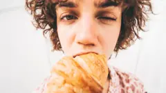 Kobieta gryzie rogalika, mrużąc przy tym jedno oko. Ujęcie typu „rybie oko” uwydatnia jej twarz, rogalika i sam kęs.