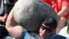 Rayno Nel, da África do Sul, visto do peito para cima, usando camiseta azul, boné preto e óculos escuros, realiza o levantamento de pedra até o ombro no World’s Strongest Man 2025, em Sacramento. Ele demonstra concentração e esforço no rosto enquanto flexiona seu enorme bíceps e levanta o braço direito para equilibrar uma enorme pedra cinza apoiada no pescoço e encostada na cabeça. Ao fundo, parcialmente visíveis, estão duas pessoas desfocadas, uma delas aplaudindo