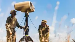 Dwóch mężczyzn w wojskowych mundurach khaki używa zestawu, który znajduje się na stojaku