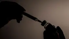 A imagem mostra, em contraluz, uma pessoa introduzindo uma seringa em um frasco de medicamento para aspirar seu conteúdo. Apenas as silhuetas das mãos, da seringa e do frasco são visíveis, sobre um fundo neutro, destacando o procedimento de preparação de uma aplicação injetável.