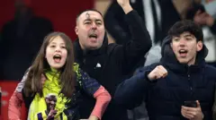 Fener taraftarı bir kız ve babası tribünlerde seviinyor. Yanlarında bir de siyah montlu genç erkek var.