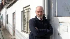 Alfonso Mateo-Sagasta, con una barba blanca corta, viste una chaqueta deportiva azul oscura, bajo la cual se ve el cuello de una camisa blanca. A su izquierda hay una casa blanca de aspecto humilde sobre cuyo muro se apoya.
