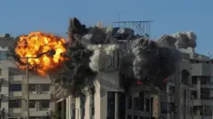 青空の下、クリーム色のタイル張りの建物が爆発し、オレンジ色の炎と黒煙が上がっている