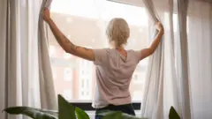 Młoda kobieta widziana od tyłu otwiera białe zasłony przed oknem 