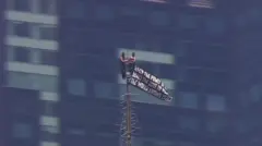 Two small figuress in black at the very top of the spire, with windows a building behind them, a black flag with white script waves to one side