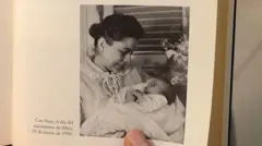 Foto em um livro mostra Naty Fernández com um vestido e com os cabelos presos para trás, admirando sua bebê, envolta em um xale branco. A legenda da foto diz, em espanhol: 'Com Naty, no dia de nascimento de Alina, 19 de março de 1956.' O nome do livro é 'Alina: Memórias da Filha Rebelde de Fidel Castro', em tradução livre
