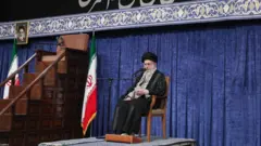علی خامنه‌ای، روی صندلی و در کنار پرچم جمهوری اسلامی ایران و منبر