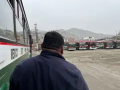 Toño, un hombre con una chaqueta azul, está parado al lado de un bus en un gran lote. Hay más buses al fondo del lote y detrás, se ve una montaña con barrios de invasión.