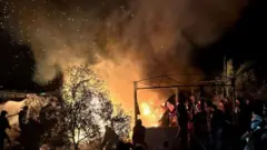 يتصاعد الدخان ليلاً بينما يقف الناس في مخيم للنازحين، بعد هجوم إسرائيلي، على خان يونس، جنوب قطاع غزة. وألسنة اللهب تبدو واضحة في الصورة.