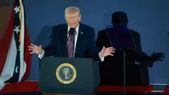 دونالد ترامپ پشت میکرفون در حال سخنرانی در حالی که دستانش را به دو طرف باز کرده، سایه او بر دیوار افتاده است