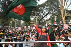 Uma multidão na rua usando faixas na cabeça com a bandeira de Banglsdesh, verde e tem um círculo vermelho.
