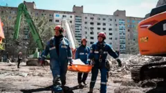 Рятувальники несуть ноші з тілом людини на фоні руїн будинку