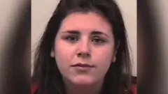 Uma mulher de cabelos longos e escuros, vestindo uma blusa vermelha, está posicionada contra um fundo liso de cor clara.