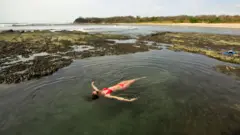 Uma mulher de biquíni vermelho relaxa em uma piscina natural. A praia é visível ao fundo