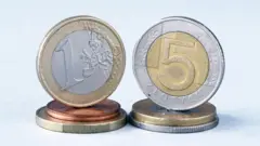 A képen egy ötzlotys és egy egyeurós érme látható egymás mellett, más aprópénzekre állítva.
