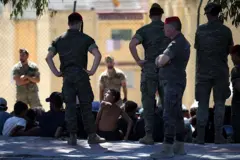Um grupo de pessoas sentadas no chão ao ar livre. Ao redor delas, um grupo de militares espanhóis em pé as vigia. Entre as pessoas sentadas, uma criança se vira e ergue o olhar para um dos militares.