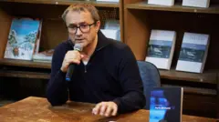 Режиссер Андрей Звягинцев в очках с микрофоном в руках сидит за коричневым столом и отвечает на вопрос на фоне стеллажей с книгами