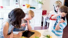Quatro irmãos juntos em uma cozinha; duas meninas olham para um tablet e dois meninos brigam por um celular