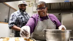 Lenore Lindsey coloca bolas de massa de biscoito sobre uma bandeja. Vamarr Hunter passa por trás dela, na cozinha da padaria dirigida por eles.