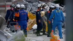 白いヘルメットとマスクを着け、青系の作業服を着た警官が、がれきのそばに立っている