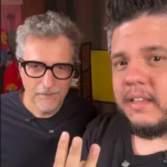 O diretor Kleber Mendonça Filho (E) e o comunicador Leo Paiva olham para câmera de celular, ambos com camiseta preta e microfones pendurados