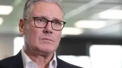 英國工黨黨魁施紀賢爵士（Sir Keir Starmer）身穿白色開領襯衫及深色西裝外套，佩戴眼鏡，神情嚴肅。他身處倫敦南部的AFC溫布頓足球俱樂部一個場地內，背景中人物經過鏡頭焦距模糊處理。（9/5/2026）