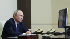 Vladimir Putin está sentado à esquerda da imagem, de perfil, com as mãos apoiadas sobre a mesa, olhando para a tela de um computador à direita da imagem, enquanto preside uma videoconferência com membros do Conselho de Segurança da Rússia.