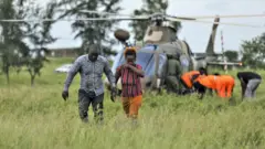 Um homem e uma mulher afastam-se de um helicóptero militar enquanto membros de uma equipe de resgate com casacos laranja ajudam alguém numa maca em Moçambique
