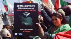 Mulher em um protesto de rua, enrolada em uma bandeira do Irã, segura um cartaz dizendo: '90 milhões de vozes, 216+ horas offline, silenciadas pelo blackout do CGRI', com outras pessoas com bandeiras iranianas ao fundo