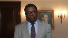 Joseph Malanji yambaye amarori imbere y'umufotozi inyuma yiwe hari ikadere