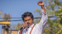 Fri for Sat - Anbo social media Vijay Sat for Sun - Profile of Indian model who met debut in Met Gala Vijay is seen during a campaign rally, wearing a white shirt with a red and yellow lanyard around his neck. He is talking into a mic in his left hand while raising his right fist.