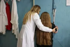Uma médica, de costas para a câmera, ajuda uma menina a subir na balança