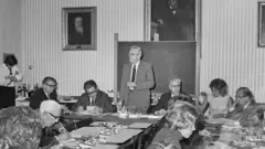 Hans Asperger está em pé em uma conferência, vestindo um terno claro e cercado por outros participantes. A foto é em preto e branco