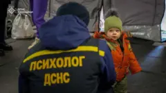 Человек в куртке с надписью "Психолог ДСНС" на спине присел на корточки перед ребенком в зимней одежде