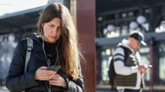 На заднем плане человек в серой кепке, темно-зеленой жилетке, белом худи и рюкзаком за плечами смотрит в телефон. На переднем плане девушка с длинными русыми волосами, проводными наушниками, в темно-синей куртке. Девушка смотрит в телефон.