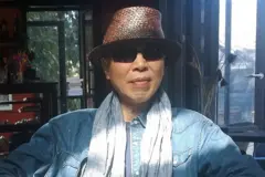 Um homem, usando chapéu marrom de palha, óculos escuros, camisa jeans e um cachecol cinza, está sentado em um ambiente interno