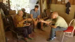 Três homens conversando sentados em ambiente doméstico; Cleo segura um gravador