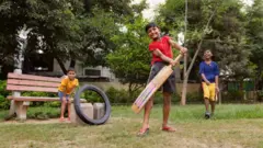 දරුවන් තිදෙනෙක් ක්‍රිකට් සෙල්ලම් කරමින් සිනාසෙමින් සිටිති.