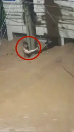 බ්‍රසීලයේ ගංවතුර මැද කාන්තාවක් විදුලි කණුවක එල්ලී දිවි බේරා ගැනීමට උත්සාහ කරන අයුරු දැක්වේ.