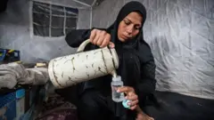 تصویری از یک فلسطینی که دریک خیمه نشته و در چوشک فرزندش از یک فلاسک آب میریزد.