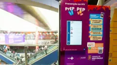 A imagem mostra uma máquina de autoatendimento para retirada de PrEP e PEP instalada em uma estação de metrô de São Paulo, ao lado de escadas rolantes movimentadas por passageiros. O equipamento, identificado com as marcas do SUS e da Prefeitura de São Paulo, possui uma tela digital e orienta os usuários a acessar o aplicativo e-saúdeSP, gerar um QR Code e utilizá-lo para retirar gratuitamente os medicamentos de prevenção ao HIV.