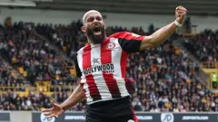 Bryan Mbeumo, wearing a red and white striped Brentford shirt, leaps and punches the air, screaming in celebration, with fans sitting in the background in a stand with yellow seats and railings